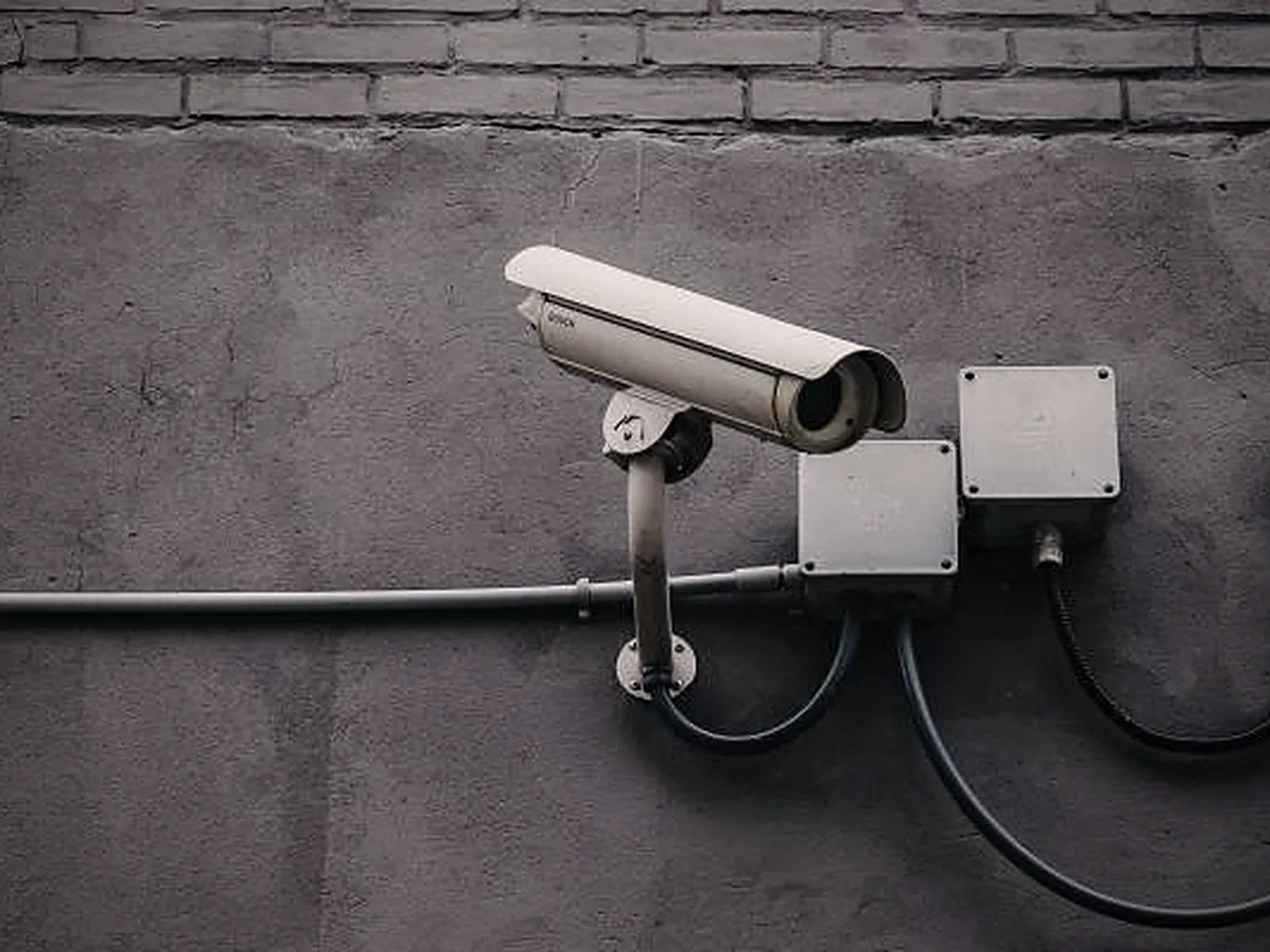A single surveillance camera bolted to a grey concrete wall beneath a brick course, wired into two junction boxes