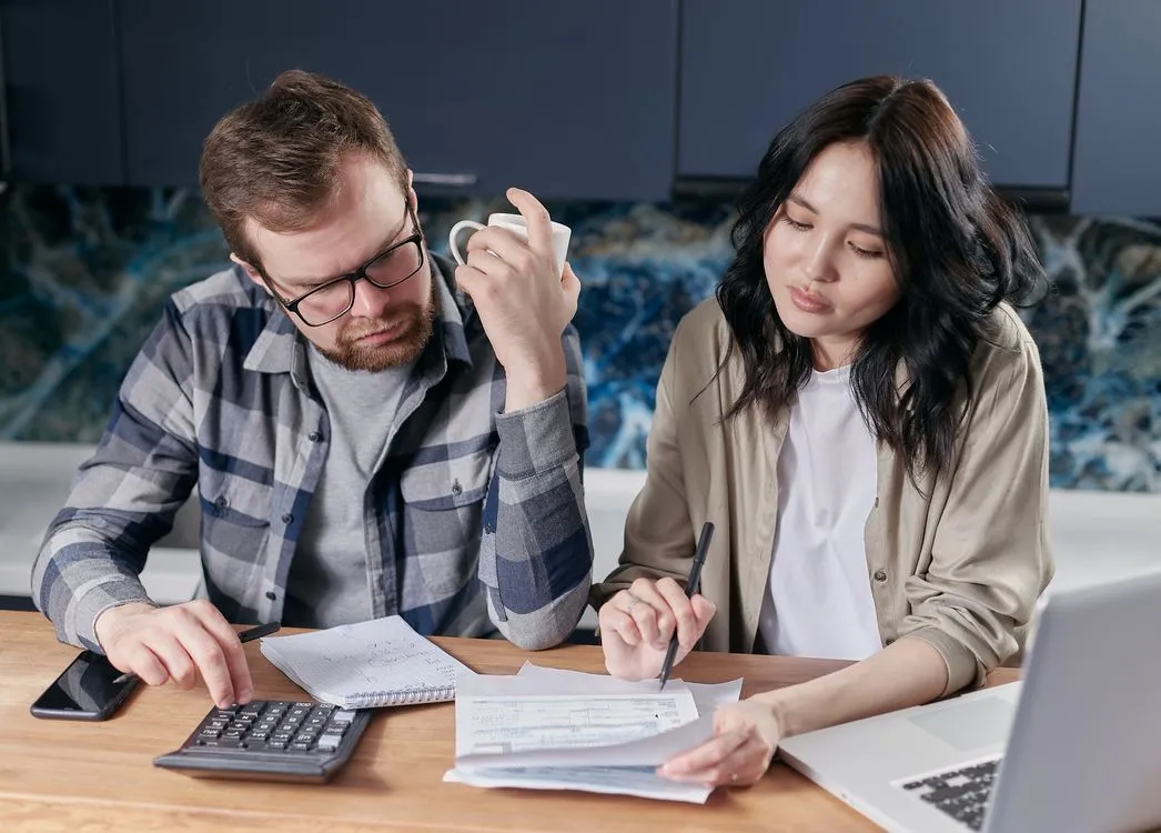 Two people at a kitchen table with laptops open to banking apps and a shared notebook