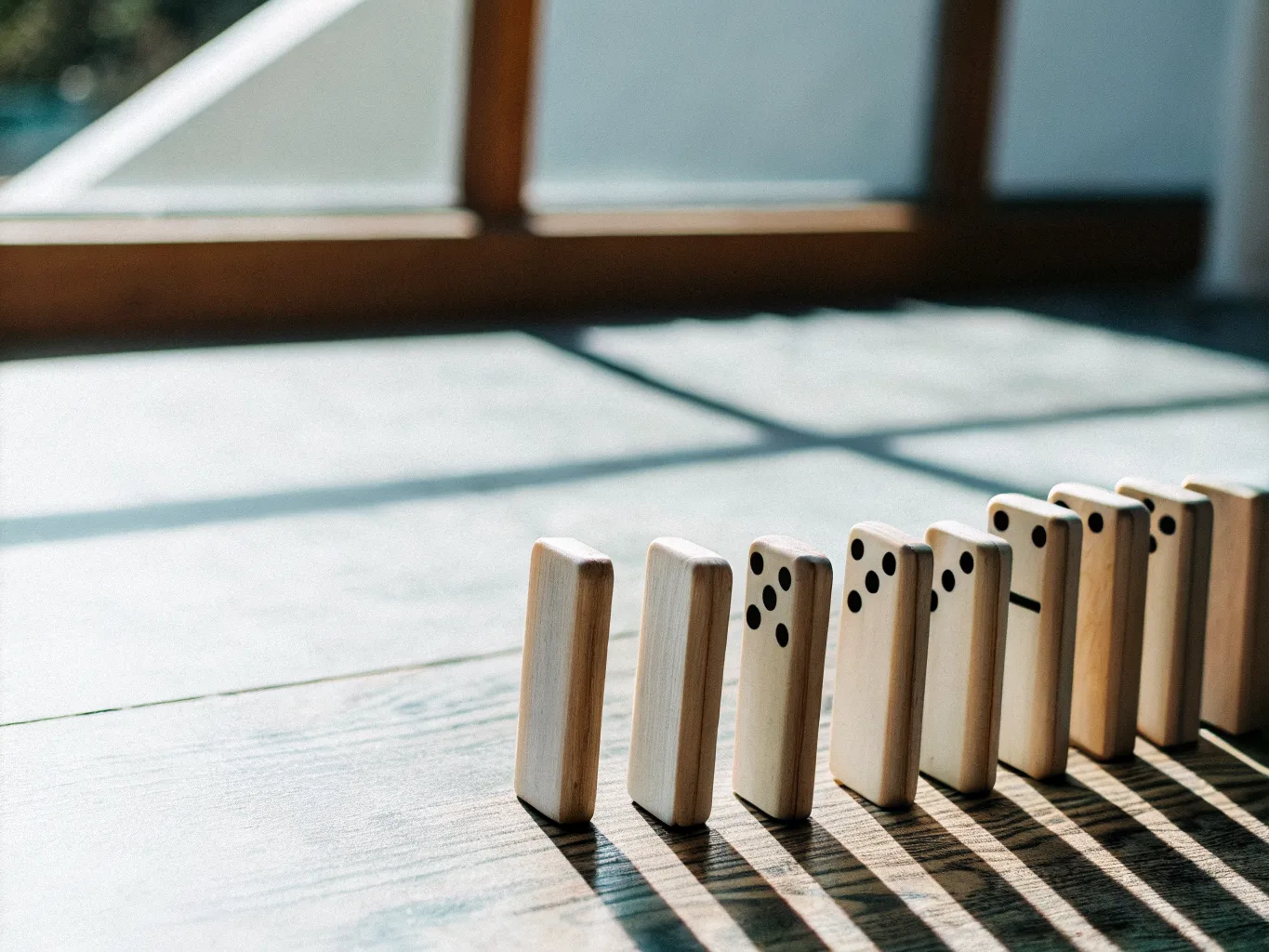 A row of blank wooden dominoes standing on a table, the first few toppling into each other, casting long shadows in low light